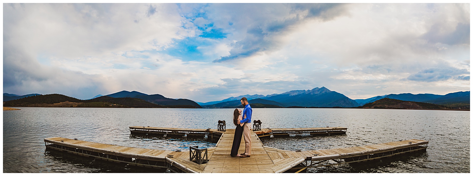 a couple stands on a dock with a lake and the mountains behind them
