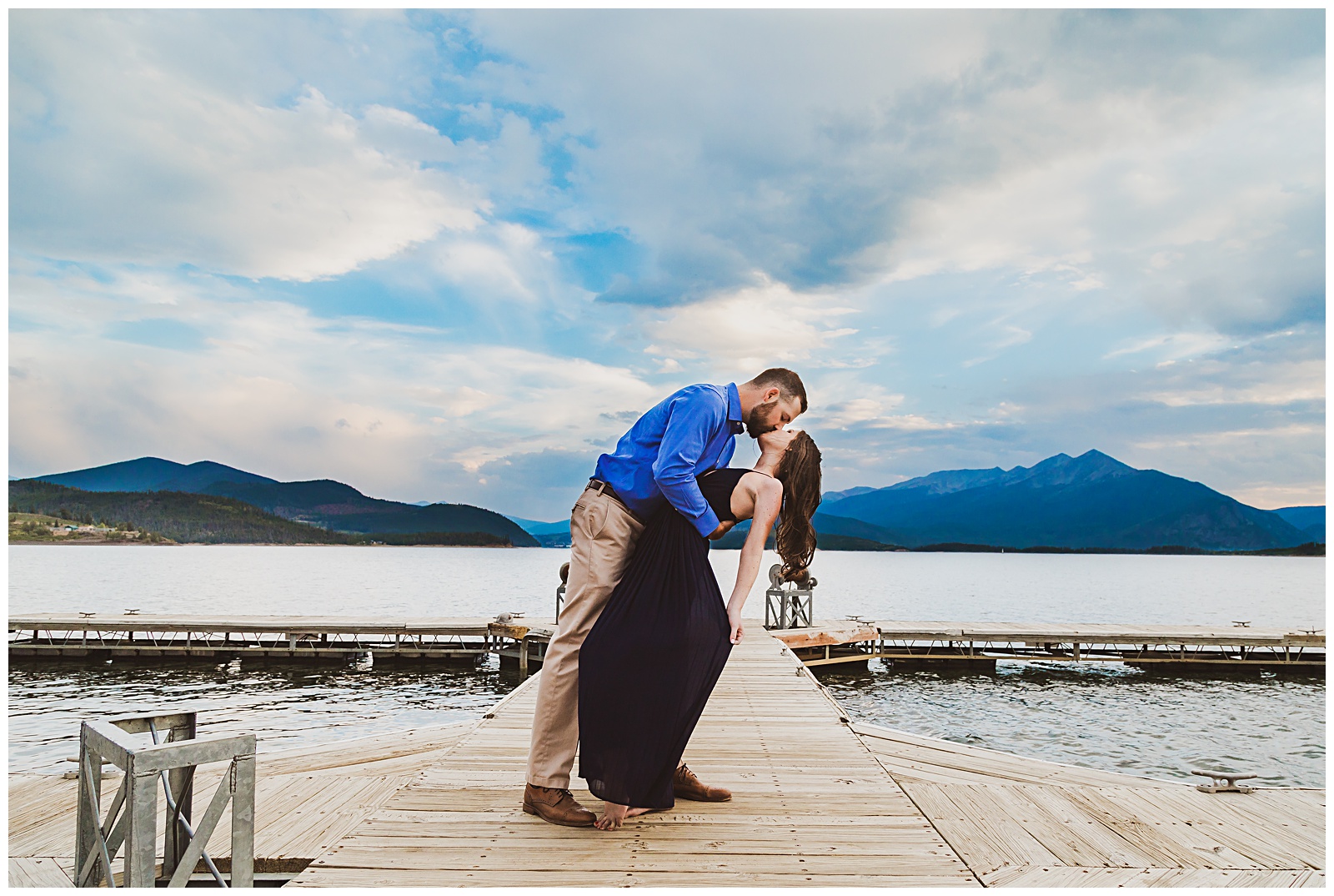 a couple kissing on a dock with a lake and the mountains behind them